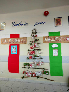 Dekoracja szkolna przedstawiająca kalendarz adwentowy ułożony w kształt choinki z naturalnych gałązek. Do gałązek przywiązane są kolorowe paczuszki oznaczone numerami dni grudnia. Po bokach wiszą duże pionowe pasy materiału, czerwony z lewej i zielony z prawej. Nad całością widnieje ręcznie wykonany napis „Kalendarz Adwentowy”.