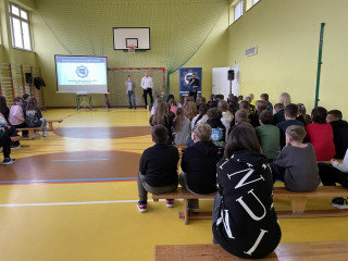 Zebrani na sali gimnastycznej w szkole uczniowie i nauczyciele siedzą na ławkach i słuchają prelekcji dwóch, stojących na końcu sali, pośrodku, policjantów z Centralnego Biura Zwalczania Cyberprzestępczości ubranych po cywilnemu. Po lewej stronie widać ekran z prezentacją. Po prawej stronie stoi dyrektor szkoły.