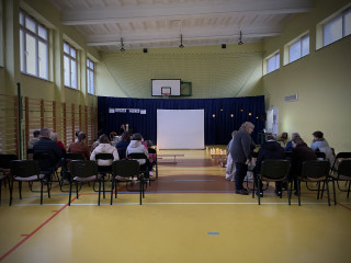 Sala gimnastyczna w szkole. Na ławkach po lewej i prawej stronie siedzą widzowie. Na końcu sali po lewej stronie stoi stolik z dwoma świecznikami, w których palą się świece, na środku sali duży, biały ekran. Na żółtej podłodze po dwóch stronach ekranu stoją zapalone świece. W tle wisi granatowa kotara.