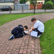 Uczeń klęczy na chodniku i pisze na karcie pracy, drugi chłopiec siedzi na trawie i pomaga koledze w pracy.