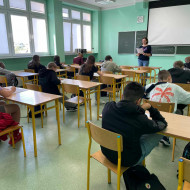 W klasie uczniowie siedzą w ławkach i piszą tekst dyktanda czytanego przez nauczycielkę.
