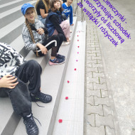 Uczennice siedzą na schodach przed szkołą, a na stopniu poniżej jest ułożona oś liczbowa ze wstążek i różyczek. 