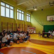Uczniowie zgromadzeni w sali gimnastycznej siedzą na ławkach i słuchają wierszy Kochanowskiego czytanych przez kolegów.