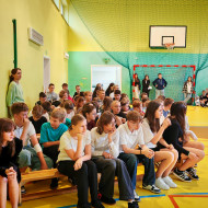 Uczniowie siedzą na ławeczkach i słuchają wierszy Jana Kochanowskiego czytanych przez młodych artystów.