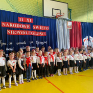 Przedszkolaki stoją przed dekoracją na sali gimnastycznej z okazji Narodowego Święta Niepodległości.
