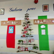 Dekoracja szkolna przedstawiająca kalendarz adwentowy ułożony w kształt choinki z naturalnych gałązek. Do gałązek przywiązane są kolorowe paczuszki oznaczone numerami dni grudnia. Po bokach wiszą duże pionowe pasy materiału, czerwony z lewej i zielony z prawej. Nad całością widnieje ręcznie wykonany napis „Kalendarz Adwentowy”.