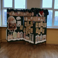 Szkolna dekoracja na której znajduje się duży napis „Kalendarz Adwentowy”, a poniżej przyczepione są małe, brązowe koperty oznaczone numerami dni. Z lewej strony stoi duża postać Mikołaja, a u dołu widoczne są ilustracje kolorowych kamieniczek pokrytych śniegiem. Całość otacza świąteczna girlanda.