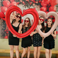 Uczennice trzymają balony w kształcie serc i pozują przed walentynkową ścianką.