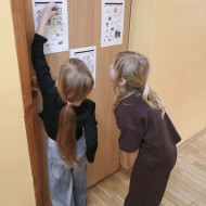 Dwie uczennice rozwiązują zadania ortograficzne wywieszone na korytarzu z okazji TKJ.