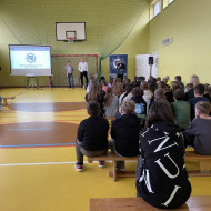 Zebrani na sali gimnastycznej w szkole uczniowie i nauczyciele siedzą na ławkach i słuchają prelekcji dwóch, stojących na końcu sali, pośrodku, policjantów z Centralnego Biura Zwalczania Cyberprzestępczości ubranych po cywilnemu. Po lewej stronie widać ekran z prezentacją. Po prawej stronie stoi dyrektor szkoły.
