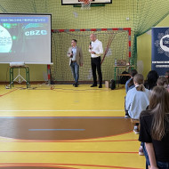 Zebrani na sali gimnastycznej w szkole uczniowie i nauczyciele siedzą na ławkach i słuchają prelekcji dwóch, stojących na końcu sali, pośrodku, policjantów z Centralnego Biura Zwalczania Cyberprzestępczości ubranych po cywilnemu. Po lewej stronie widać ekran z prezentacją.