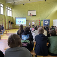 Zebrani na sali gimnastycznej w szkole uczniowie i nauczyciele siedzą na ławkach i słuchają prelekcji dwóch, stojących na końcu sali, pośrodku, policjantów z Centralnego Biura Zwalczania Cyberprzestępczości ubranych po cywilnemu. Po lewej stronie widać ekran z prezentacją.