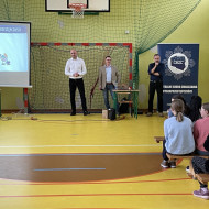 Zebrani na sali gimnastycznej w szkole uczniowie i nauczyciele siedzą na ławkach i słuchają prelekcji dwóch, stojących na końcu sali, pośrodku, policjantów z Centralnego Biura Zwalczania Cyberprzestępczości ubranych po cywilnemu. Po lewej stronie widać ekran z prezentacją. Po prawej stronie stoi dyrektor szkoły.
