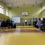 Zebrani na sali gimnastycznej w szkole uczniowie i nauczyciele siedzą na ławkach i słuchają prelekcji dwóch, stojących na końcu sali, pośrodku, policjantów z Centralnego Biura Zwalczania Cyberprzestępczości ubranych po cywilnemu. Po lewej stronie widać ekran z prezentacją.