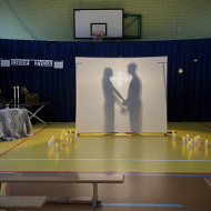 Sala gimnastyczna w szkole. Na końcu sali po lewej stronie stoi stolik z dwoma świecznikami, w których palą się świece, przy którym siedzi czytający wiersze uczeń. Na środku sali duży, biały ekran. Na żółtej podłodze po dwóch stronach ekranu stoją zapalone świece. W tle wisi granatowa kotara. Na ekranie wyświetlają się cienie dwóch postaci - chłopca i dziewczynki, trzymających się za ręce.