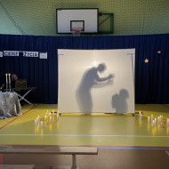 Sala gimnastyczna w szkole. Na końcu sali po lewej stronie stoi stolik z dwoma świecznikami, w których palą się świece, przy którym siedzi czytający wiersze uczeń. Na środku sali duży, biały ekran. Na żółtej podłodze po dwóch stronach ekranu stoją zapalone świece. W tle wisi granatowa kotara. Na ekranie wyświetlają się cienie dwóch postaci - po prawej skulonego dziecka, po lewej mężczyzny majaczącego rękoma.