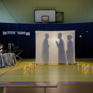 Sala gimnastyczna w szkole. Na końcu sali po lewej stronie stoi stolik z dwoma świecznikami, w których palą się świece, przy którym siedzi czytający wiersze uczeń. Na środku sali duży, biały ekran. Na żółtej podłodze po dwóch stronach ekranu stoją zapalone świece. W tle wisi granatowa kotara. Na ekranie wyświetlają się cienie trzech stojących postaci - od lewej mężczyzny, księdza i mężczyzny z bujną, kręconą fryzurą.