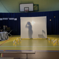 Sala gimnastyczna w szkole. Na końcu sali po lewej stronie stoi stolik z dwoma świecznikami, w których palą się świece, przy którym siedzi czytający wiersze uczeń. Na środku sali duży, biały ekran. Na żółtej podłodze po dwóch stronach ekranu stoją zapalone świece. W tle wisi granatowa kotara. Na ekranie po lewej stronie wyświetla się cień kobiecej postaci trzymającej się za głowę, poi prawej stojące krzesło.