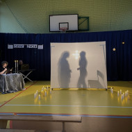 Sala gimnastyczna w szkole. Na końcu sali po lewej stronie stoi stolik z dwoma świecznikami, w których palą się świece, przy którym siedzi czytający wiersze uczeń. Na środku sali duży, biały ekran. Na żółtej podłodze po dwóch stronach ekranu stoją zapalone świece. W tle wisi granatowa kotara. Na ekranie po lewej stronie wyświetlają się cienie dwóch kobiecych postaci skierowanych twarzami do siebie, po prawej stojące krzesło.