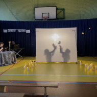 Sala gimnastyczna w szkole. Na końcu sali po lewej stronie stoi stolik z dwoma świecznikami, w których palą się świece, przy którym siedzi czytający wiersze uczeń. Na środku sali duży, biały ekran. Na żółtej podłodze po dwóch stronach ekranu stoją zapalone świece. W tle wisi granatowa kotara. Na ekranie wyświetlają się cienie dwóch dziewczynek podrzucających maskotki.