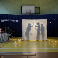 Sala gimnastyczna w szkole. Na końcu sali po lewej stronie stoi stolik z dwoma świecznikami, w których palą się świece, przy którym siedzi czytający wiersze uczeń. Na środku sali duży, biały ekran. Na żółtej podłodze po dwóch stronach ekranu stoją zapalone świece. W tle wisi granatowa kotara. Na ekranie wyświetlają się cienie trzech osób agresywnie machających do siebie rękami.