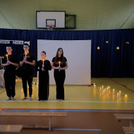 Sala gimnastyczna w szkole. Na środku sali duży, biały ekran. Na żółtej podłodze po dwóch stronach ekranu stoją zapalone świece. W tle wisi granatowa kotara. Przed ekranem stoi pięcioro uczniów z zapalonymi świecami w dłoniach. Kolejny uczeń idzie po prawej stronie mówiąc wiersz do mikrofonu.