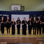 Sala gimnastyczna w szkole. Na środku sali duży, biały ekran. Na żółtej podłodze po dwóch stronach ekranu stoją zapalone świece. W tle wisi granatowa kotara. Przed ekranem stoi dziesięcioro uczniów z zapalonymi świecami w dłoniach.