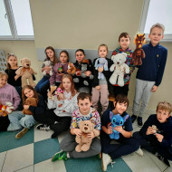 Część uczniów klasy 5 siedzi na ławce na korytarzu szkoły, część siedzi przed nimi na podłodze i przytulają swoje misie.