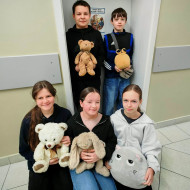 Trzy dziewczyny siedzą na podłodze, dwóch chłopców stoi za nimi w drzwiach do klasy, wszyscy trzymają maskotki.