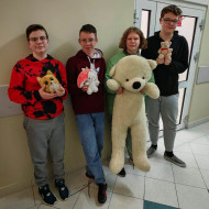 Uczniowie klasy 8 stoją na korytarzu, jeden z nich trzyma bardzo dużego białego misia, trzej pozostali mają mniejsze maskotki.