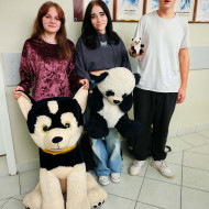 Troje uczniów stoi na korytarzu, jedna uczennica trzyma maskotkę bardzo dużego psa, druga dużego misia pandę, uczeń w ręce ma malutkiego misia pandę.