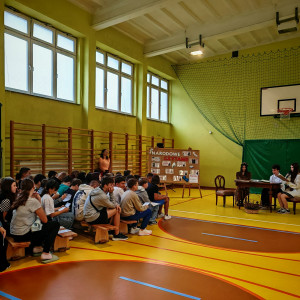 Uczniowie zgromadzeni w sali gimnastycznej siedzą na ławkach i słuchają wierszy Kochanowskiego czytanych przez kolegów.