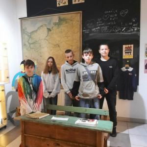 Uczniowie w dawnej klasie przy ławce szkolnej przed czarną tablicą.