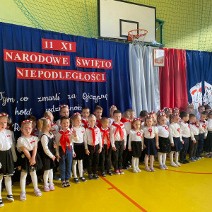 Przedszkolaki stoją przed dekoracją na sali gimnastycznej z okazji Narodowego Święta Niepodległości.