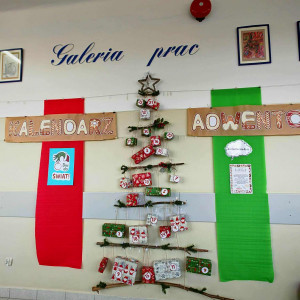 Dekoracja szkolna przedstawiająca kalendarz adwentowy ułożony w kształt choinki z naturalnych gałązek. Do gałązek przywiązane są kolorowe paczuszki oznaczone numerami dni grudnia. Po bokach wiszą duże pionowe pasy materiału, czerwony z lewej i zielony z prawej. Nad całością widnieje ręcznie wykonany napis „Kalendarz Adwentowy”.