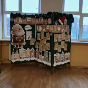 Szkolna dekoracja na której znajduje się duży napis „Kalendarz Adwentowy”, a poniżej przyczepione są małe, brązowe koperty oznaczone numerami dni. Z lewej strony stoi duża postać Mikołaja, a u dołu widoczne są ilustracje kolorowych kamieniczek pokrytych śniegiem. Całość otacza świąteczna girlanda.