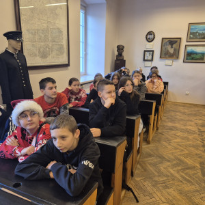 Uczniowie siedzą w zabytkowych ławkach szkolnych.