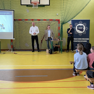 Zebrani na sali gimnastycznej w szkole uczniowie i nauczyciele siedzą na ławkach i słuchają prelekcji dwóch, stojących na końcu sali, pośrodku, policjantów z Centralnego Biura Zwalczania Cyberprzestępczości ubranych po cywilnemu. Po lewej stronie widać ekran z prezentacją. Po prawej stronie stoi dyrektor szkoły.