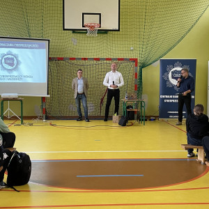 Zebrani na sali gimnastycznej w szkole uczniowie i nauczyciele siedzą na ławkach i słuchają prelekcji dwóch, stojących na końcu sali, pośrodku, policjantów z Centralnego Biura Zwalczania Cyberprzestępczości ubranych po cywilnemu. Po lewej stronie widać ekran z prezentacją. Po prawej stronie stoi dyrektor szkoły.