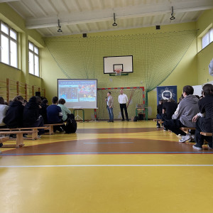 Zebrani na sali gimnastycznej w szkole uczniowie i nauczyciele siedzą na ławkach i słuchają prelekcji dwóch, stojących na końcu sali, pośrodku, policjantów z Centralnego Biura Zwalczania Cyberprzestępczości ubranych po cywilnemu. Po lewej stronie widać ekran z prezentacją.