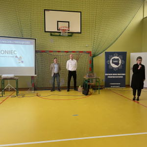 Sali gimnastyczna w szkole. Dwóch stojących pośrodku policjantów z Centralnego Biura Zwalczania Cyberprzestępczości ubranych po cywilnemu wygłasza prelekcję. Po lewej stronie widać ekran z prezentacją. Po prawej stronie stoi dyrektorka szkoły.