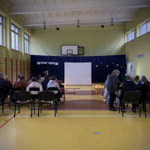 Sala gimnastyczna w szkole. Na ławkach po lewej i prawej stronie siedzą widzowie. Na końcu sali po lewej stronie stoi stolik z dwoma świecznikami, w których palą się świece, na środku sali duży, biały ekran. Na żółtej podłodze po dwóch stronach ekranu stoją zapalone świece. W tle wisi granatowa kotara.