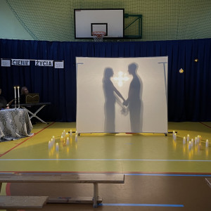 Sala gimnastyczna w szkole. Na końcu sali po lewej stronie stoi stolik z dwoma świecznikami, w których palą się świece, przy którym siedzi czytający wiersze uczeń. Na środku sali duży, biały ekran. Na żółtej podłodze po dwóch stronach ekranu stoją zapalone świece. W tle wisi granatowa kotara. Na ekranie wyświetlają się cienie dwóch postaci - chłopca i dziewczynki, trzymających się za ręce.