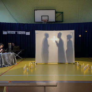 Sala gimnastyczna w szkole. Na końcu sali po lewej stronie stoi stolik z dwoma świecznikami, w których palą się świece, przy którym siedzi czytający wiersze uczeń. Na środku sali duży, biały ekran. Na żółtej podłodze po dwóch stronach ekranu stoją zapalone świece. W tle wisi granatowa kotara. Na ekranie wyświetlają się cienie trzech stojących postaci - od lewej mężczyzny, księdza i mężczyzny z bujną, kręconą fryzurą.