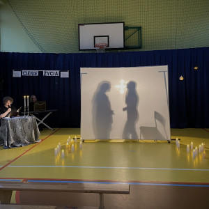 Sala gimnastyczna w szkole. Na końcu sali po lewej stronie stoi stolik z dwoma świecznikami, w których palą się świece, przy którym siedzi czytający wiersze uczeń. Na środku sali duży, biały ekran. Na żółtej podłodze po dwóch stronach ekranu stoją zapalone świece. W tle wisi granatowa kotara. Na ekranie po lewej stronie wyświetlają się cienie dwóch kobiecych postaci skierowanych twarzami do siebie, po prawej stojące krzesło.