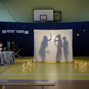 Sala gimnastyczna w szkole. Na końcu sali po lewej stronie stoi stolik z dwoma świecznikami, w których palą się świece, przy którym siedzi czytający wiersze uczeń. Na środku sali duży, biały ekran. Na żółtej podłodze po dwóch stronach ekranu stoją zapalone świece. W tle wisi granatowa kotara. Na ekranie wyświetlają się cienie trzech osób agresywnie machających do siebie rękami.