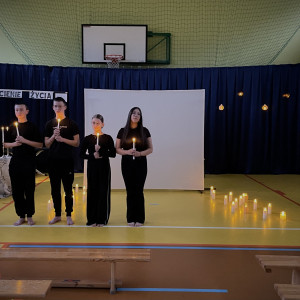 Sala gimnastyczna w szkole. Na środku sali duży, biały ekran. Na żółtej podłodze po dwóch stronach ekranu stoją zapalone świece. W tle wisi granatowa kotara. Przed ekranem stoi pięcioro uczniów z zapalonymi świecami w dłoniach. Kolejny uczeń idzie po prawej stronie mówiąc wiersz do mikrofonu.