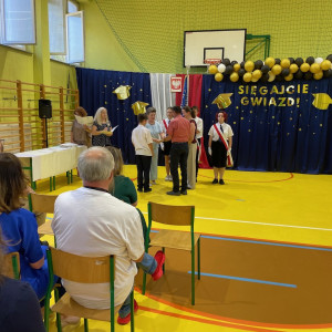 Sala gimnastyczna w szkole, po lewej stronie dyrekcja szkoły wręcza nagrodę uczniowi oraz jego rodzicom