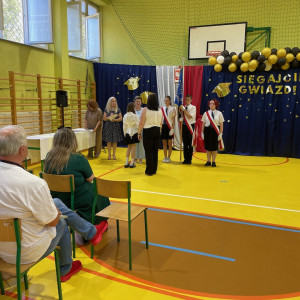 Sala gimnastyczna w szkole, po lewej stronie dyrekcja szkoły wręcza nagrodę uczennicy oraz jej rodzicom