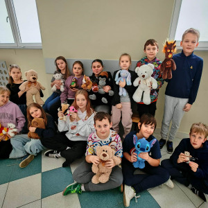 Część uczniów klasy 5 siedzi na ławce na korytarzu szkoły, część siedzi przed nimi na podłodze i przytulają swoje misie.